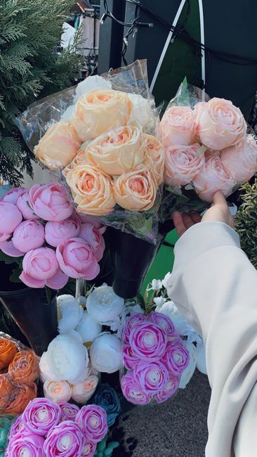 A close-up view of several bouquets of roses displayed outdoors at a flower stall. The arrangements include pale peach roses, soft pink roses, white roses, and vibrant pink roses, each wrapped in clea