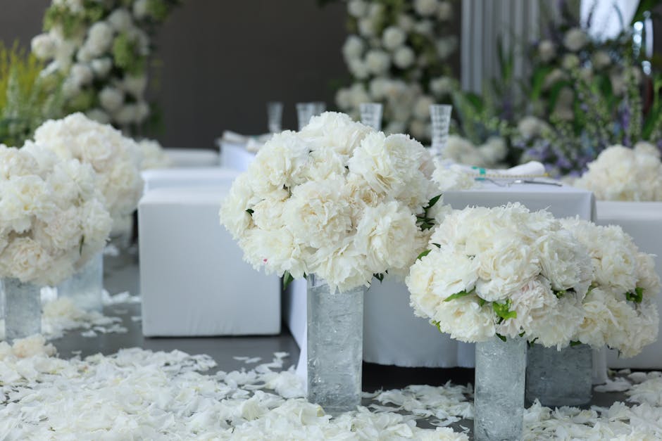 A close-up of three elegant floral arrangements featuring large, fluffy white peonies and hydrangeas in cylindrical glass vases, positioned on a surface scattered with white flower petals. The arrange