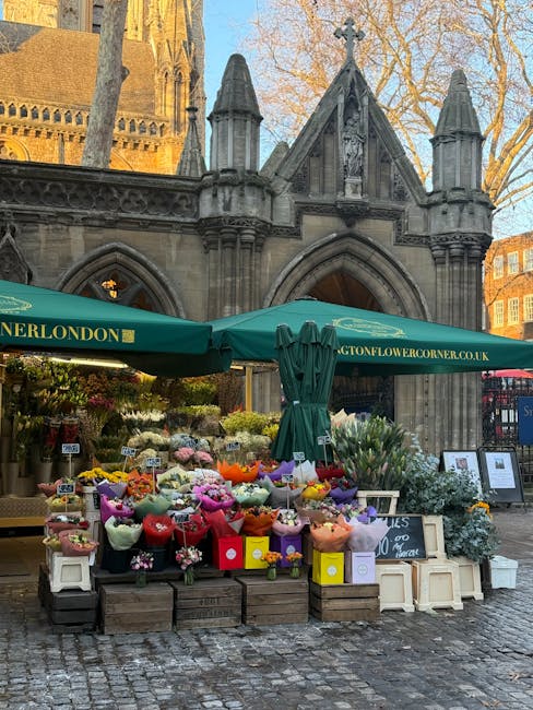 A flower stall situated outdoors in front of a historical stone building with pointed arches and decorative turrets, featuring a green canopy with the URL 'langtonflowercorner.co.uk' printed on it. Th