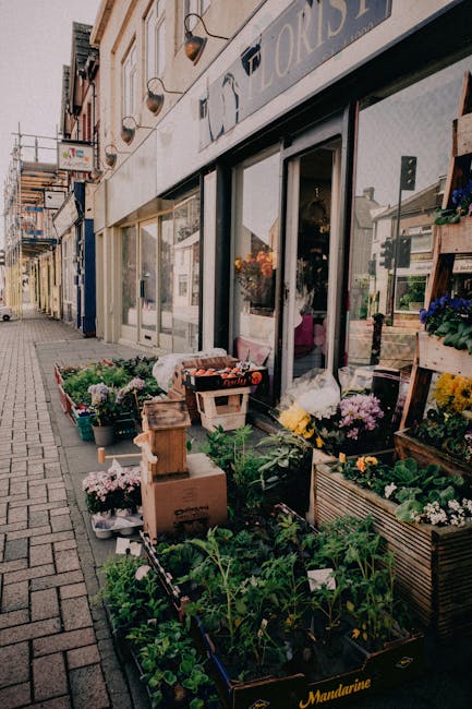A flower shop outside a building on a pedestrian street, displaying an assortment of fresh flowers and plants arranged in wooden crates, cardboard boxes, and plastic containers. The flowers include pi