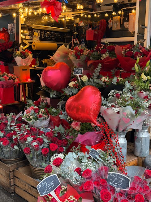 A vibrant floral display at a flower shop, featuring an assortment of fresh red roses, white and green foliage, and decorative heart-shaped balloons. The arrangement includes bouquets wrapped in red a